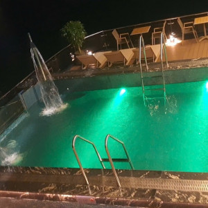 Nighttime pool with green lights, a fountain jet, and metal ladders; lounge chairs on the deck in the background.