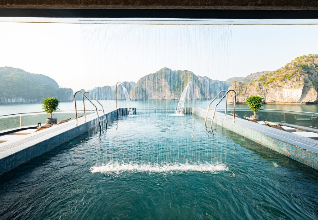 Luxury seaside pool with cascading water jets and lounge chairs, framed by rocky islands in the distance.