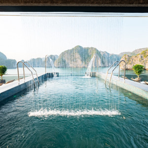 Luxury seaside pool with cascading water jets and lounge chairs, framed by rocky islands in the distance.