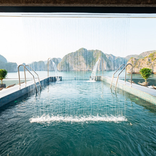 Luxury seaside pool with cascading water jets and lounge chairs, framed by rocky islands in the distance.
