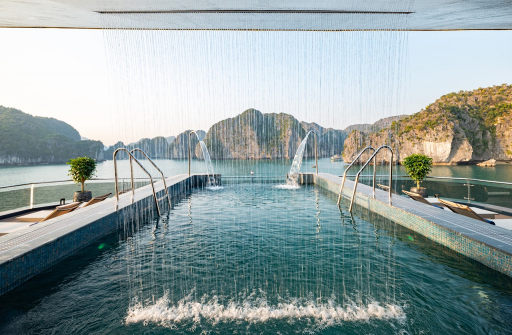 Infinity pool on a resort deck with water jets and a bay framed by rocky cliffs in the distance.