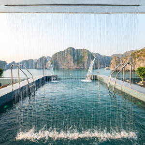 Infinity pool on a resort deck with water jets and a bay framed by rocky cliffs in the distance.
