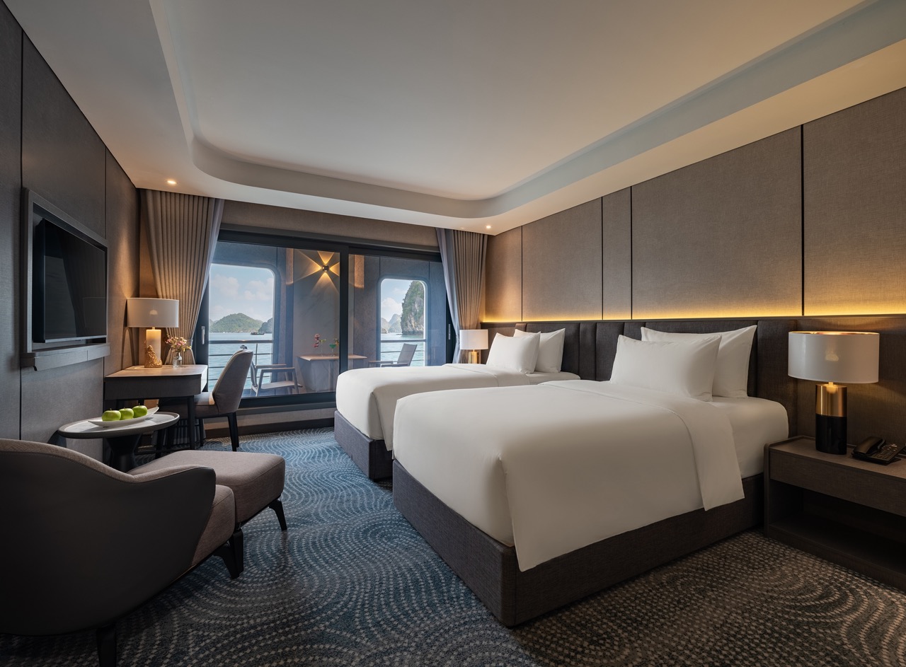 Modern hotel room with two white‑duvet beds, seating area, and balcony overlooking water, bright and inviting.