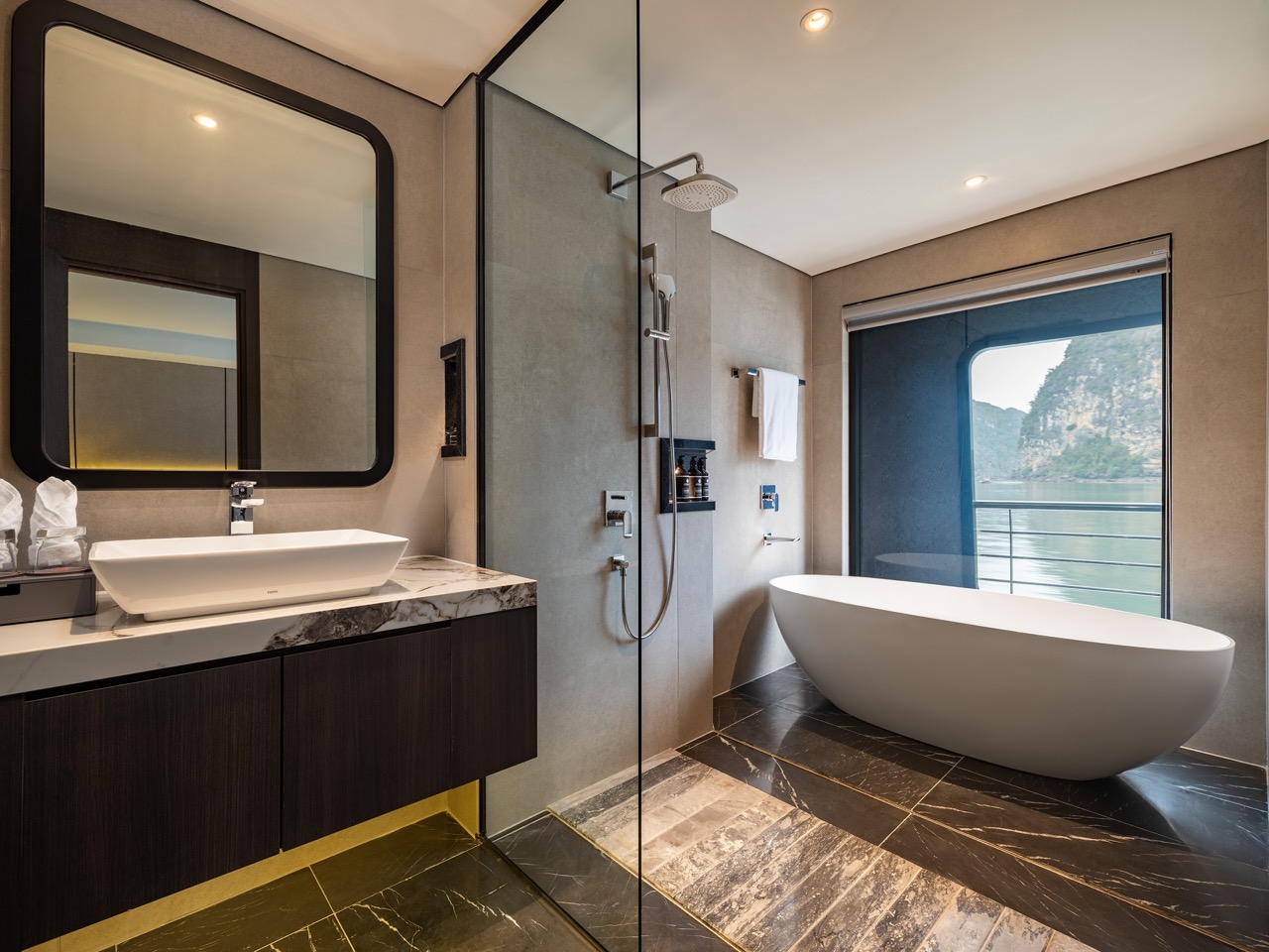 Modern bathroom with a freestanding white bathtub by a large window overlooking water and cliffs outside.