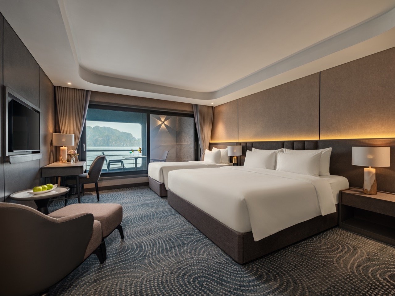 Bright hotel room with two large beds, a seating area, and a balcony overlooking mountains and water.