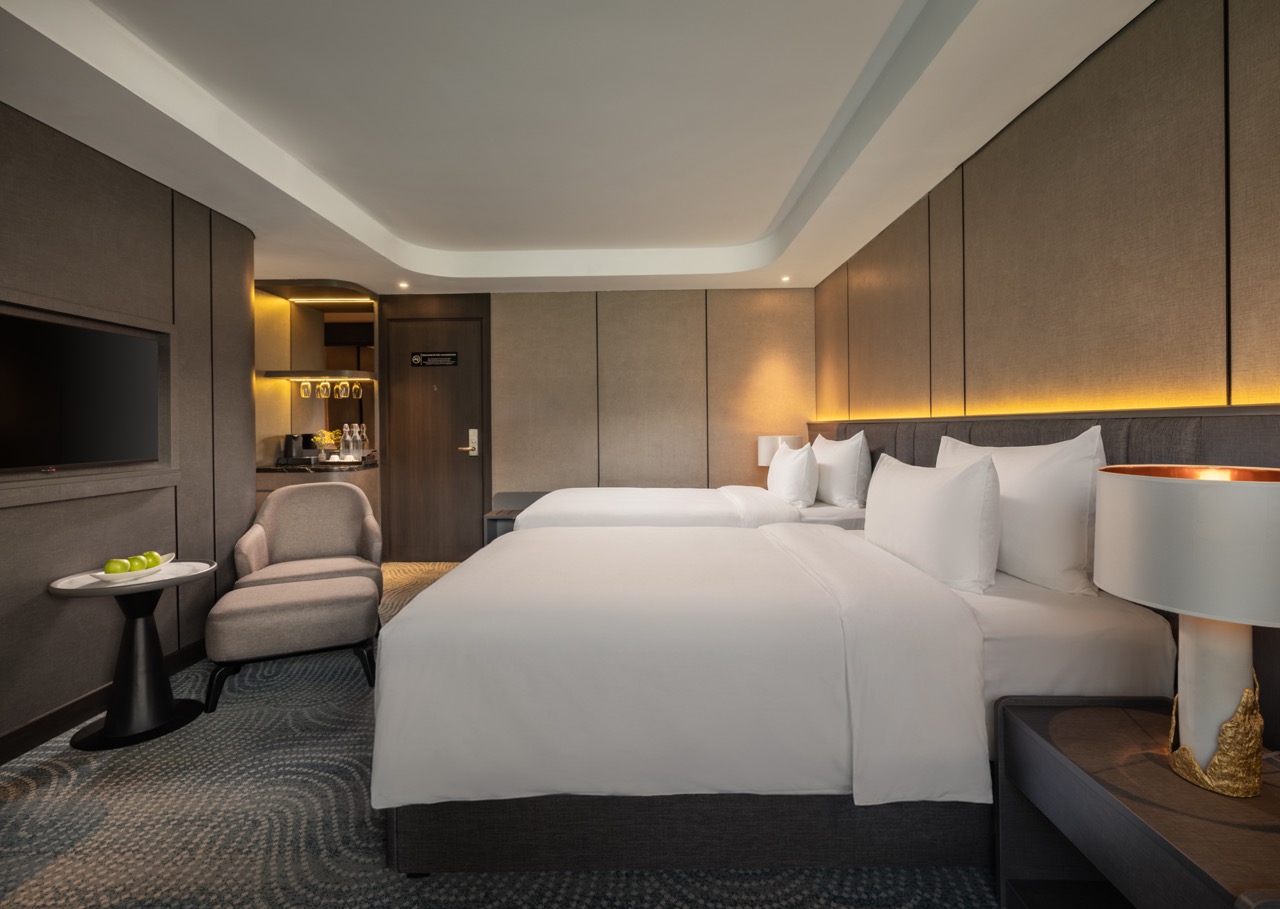 Hotel room with two neatly made beds, gray headboards, and warm ambient lighting along the wall, plus a seating area and a TV on the wall.