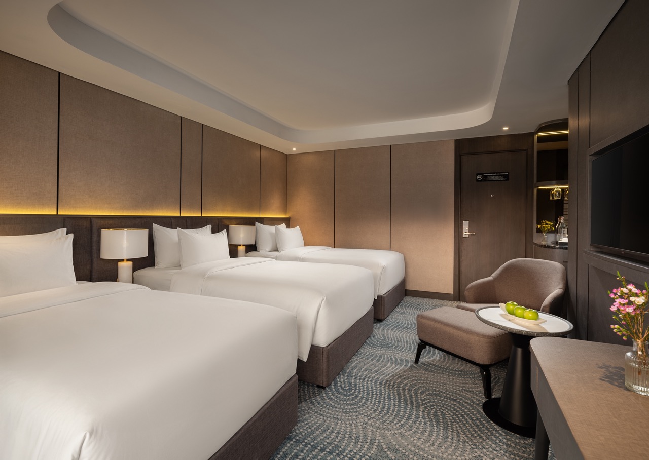 Hotel room with three white beds, beige headboards, and warm wall lighting along the length of the headboard wall.