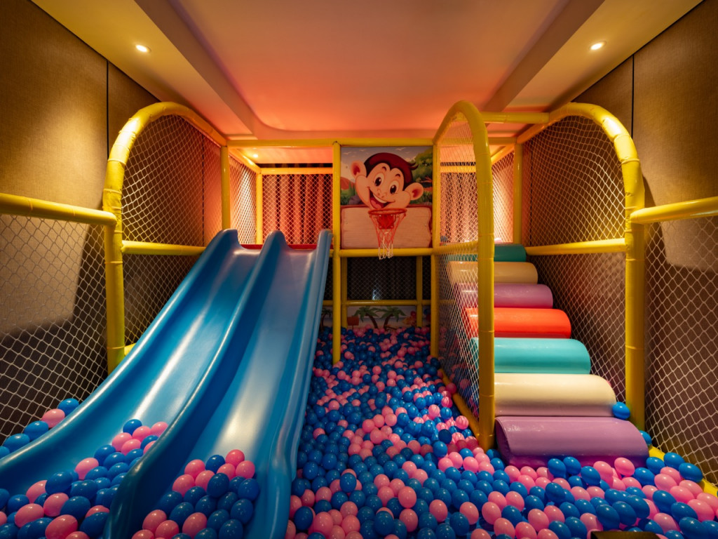 Indoor kids' play area with a blue double slide and a colorful ball pit (pink/blue balls) surrounded by yellow netted walls.