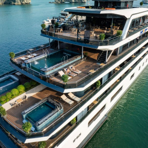 A large multi-deck luxury yacht with swimming pools and sun loungers on each level, afloat on calm blue water.