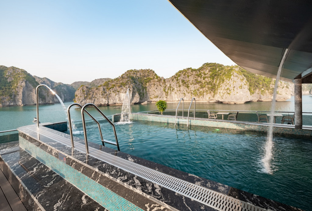Rooftop pool with metal ladders and water jets, overlooking turquoise water and rocky cliffs in the distance.