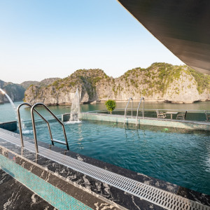 Rooftop pool with metal ladders and water jets, overlooking turquoise water and rocky cliffs in the distance.