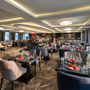 Elegant waterfront restaurant dining room with marble floors, tables set with wine glasses and red napkins under chandeliers.