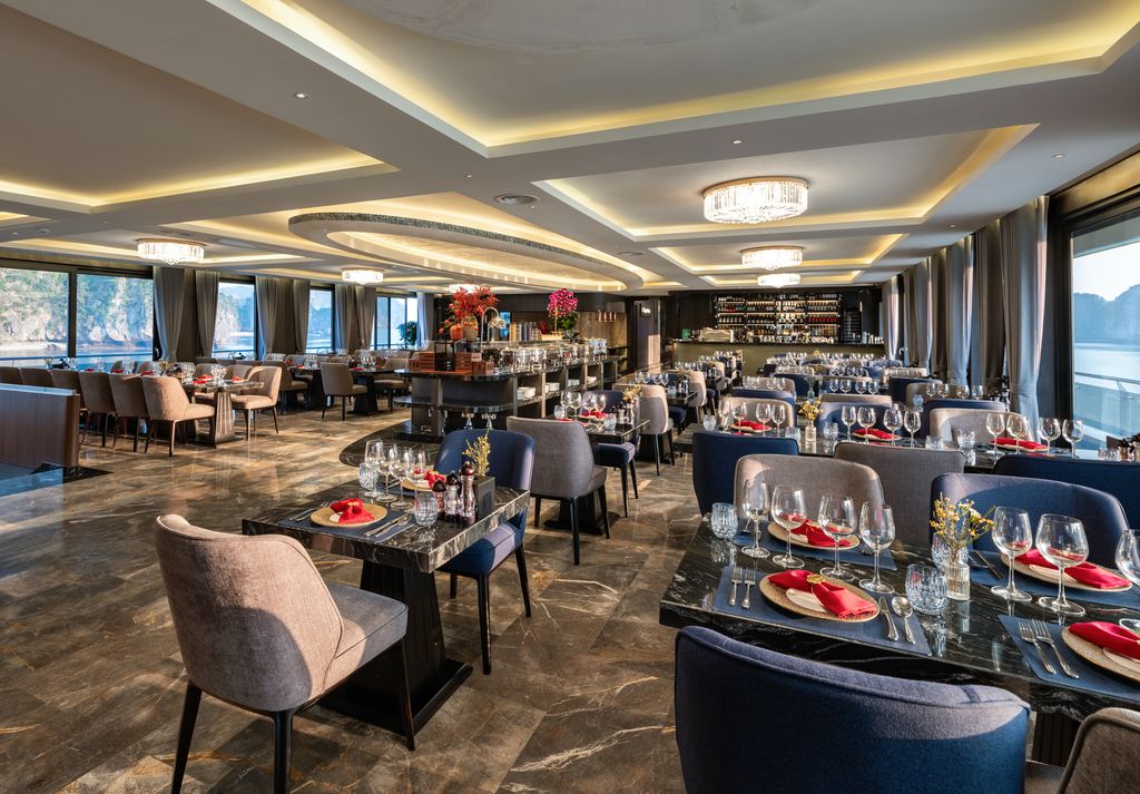 Elegant waterfront restaurant dining room with marble floors, tables set with wine glasses and red napkins under chandeliers.