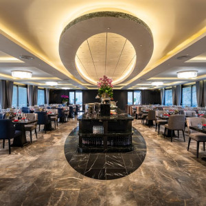 Elegant upscale restaurant dining room with a central circular ceiling feature, marble floors, and rows of tables set with wine glasses and red napkins while a centerpiece bar stands in the middle.