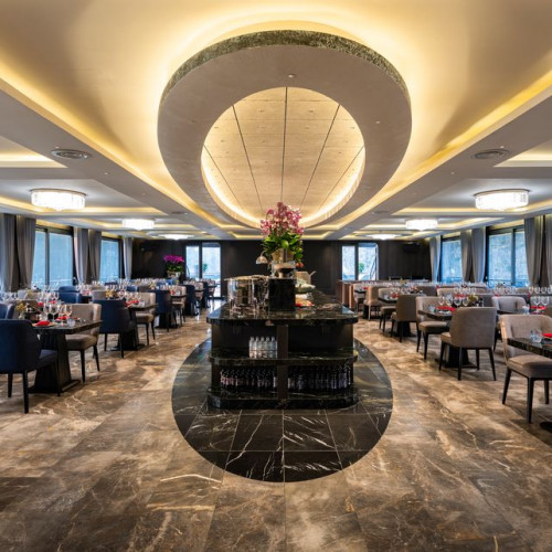 Elegant upscale restaurant dining room with a central circular ceiling feature, marble floors, and rows of tables set with wine glasses and red napkins while a centerpiece bar stands in the middle.