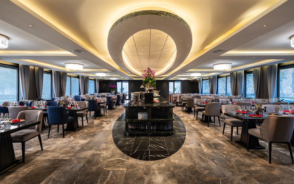 Elegant upscale restaurant dining room with a central circular ceiling feature, marble floors, and rows of tables set with wine glasses and red napkins while a centerpiece bar stands in the middle.