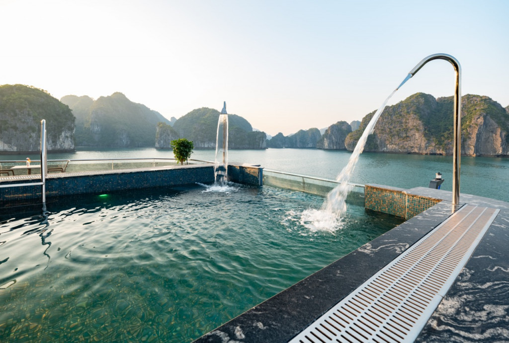Resort pool overlooking turquoise water and limestone cliffs, with a curved fountain and water spilling into the pool at sunset.