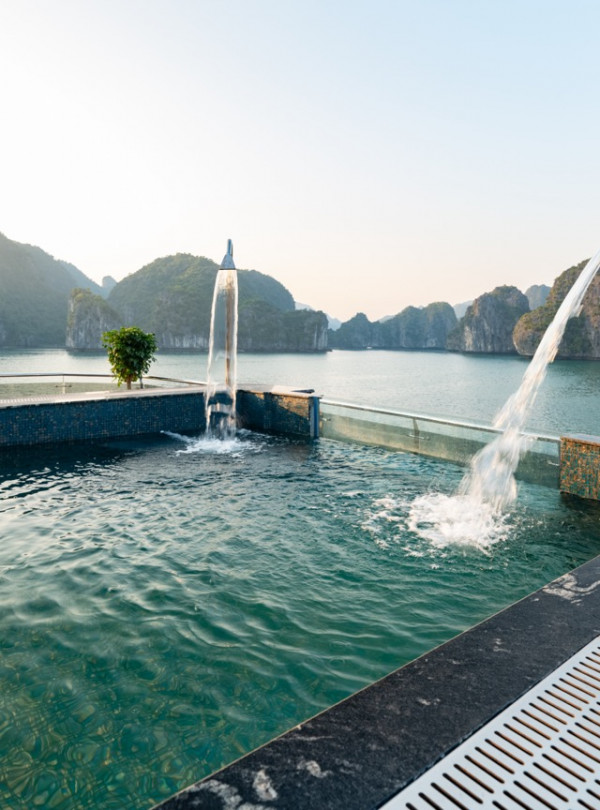 Resort pool overlooking turquoise water and limestone cliffs, with a curved fountain and water spilling into the pool at sunset.