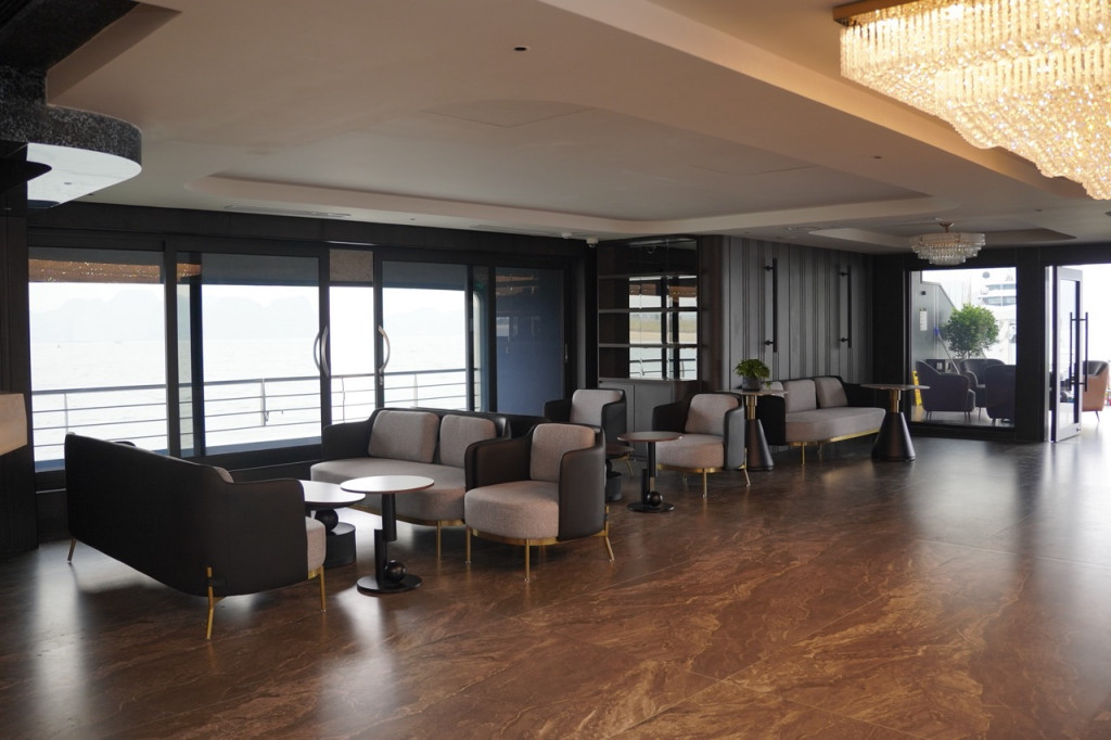 Modern lounge with gray sofas and round tables overlooking a waterfront through large glass doors; chandelier lighting overhead.