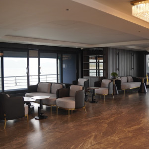 Modern lounge with gray sofas and round tables overlooking a waterfront through large glass doors; chandelier lighting overhead.
