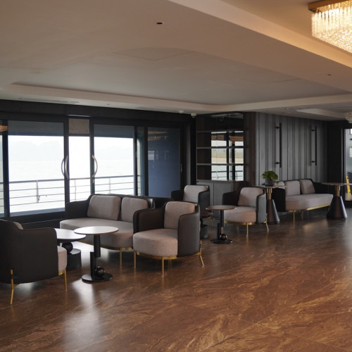 Modern lounge with gray sofas and round tables overlooking a waterfront through large glass doors; chandelier lighting overhead.