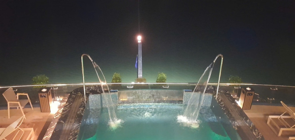 Night view of a rooftop pool with two curved water fountains arching into the pool and a central illuminated obelisk with a flag in the background.