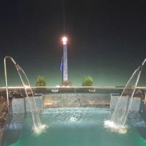 Night view of a rooftop pool with two curved water fountains arching into the pool and a central illuminated obelisk with a flag in the background.