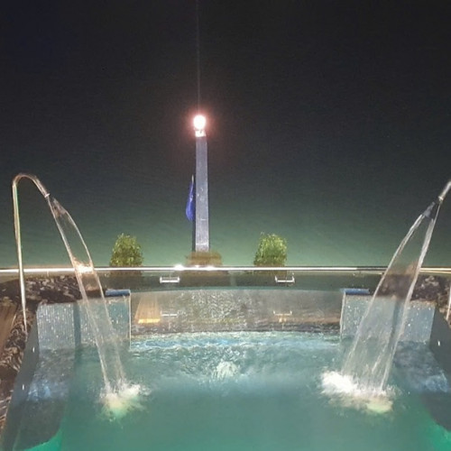 Night view of a rooftop pool with two curved water fountains arching into the pool and a central illuminated obelisk with a flag in the background.