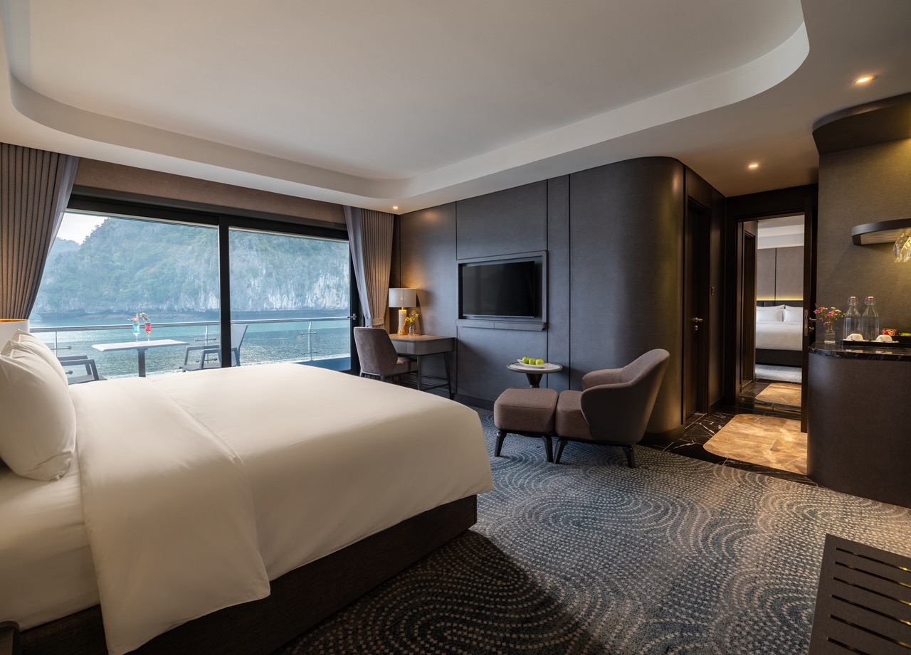 Luxurious hotel room with a king bed facing a balcony overlooking the sea, plus a seating area and wall-mounted TV.