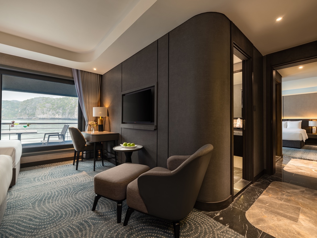 Luxurious hotel suite living area with a gray accent wall, TV, seating, and a balcony overlooking water and hills.