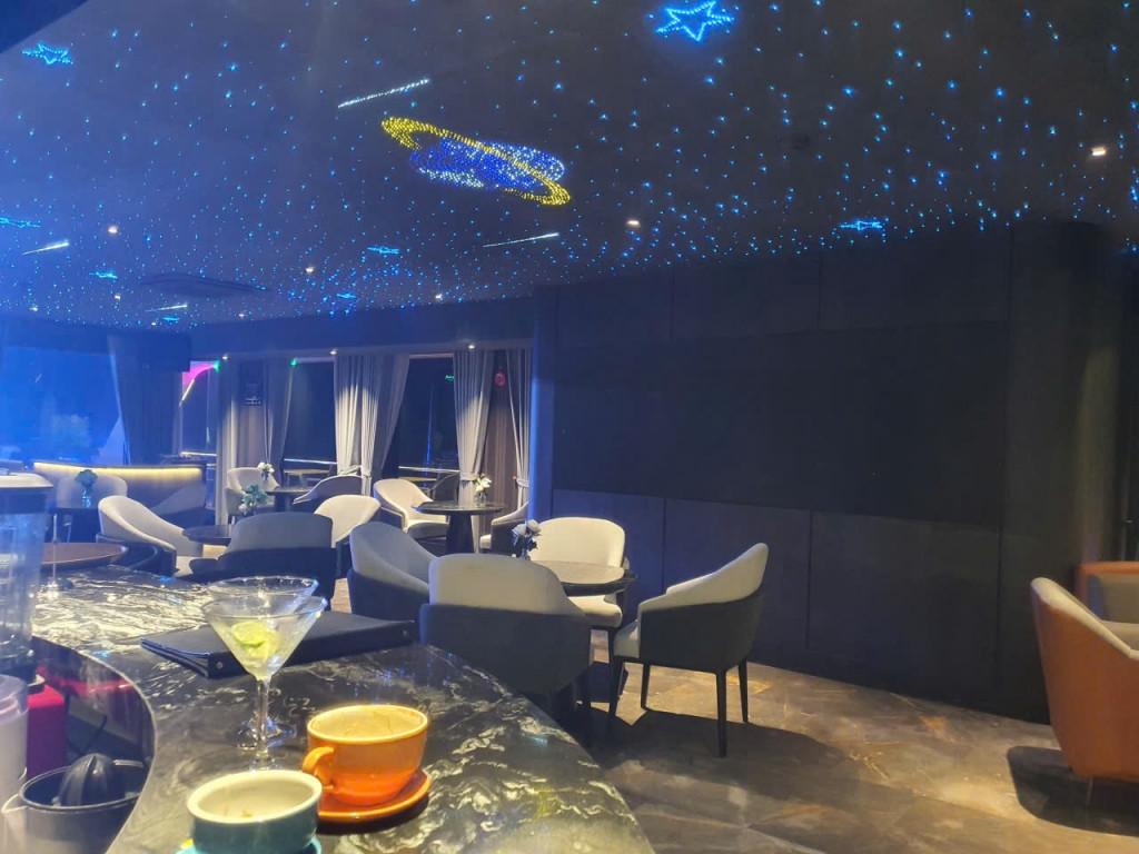 Lounge with a starry ceiling of blue lights, round tables and gray chairs; drinks on a marble bar in the foreground.