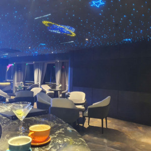 Lounge with a starry ceiling of blue lights, round tables and gray chairs; drinks on a marble bar in the foreground.