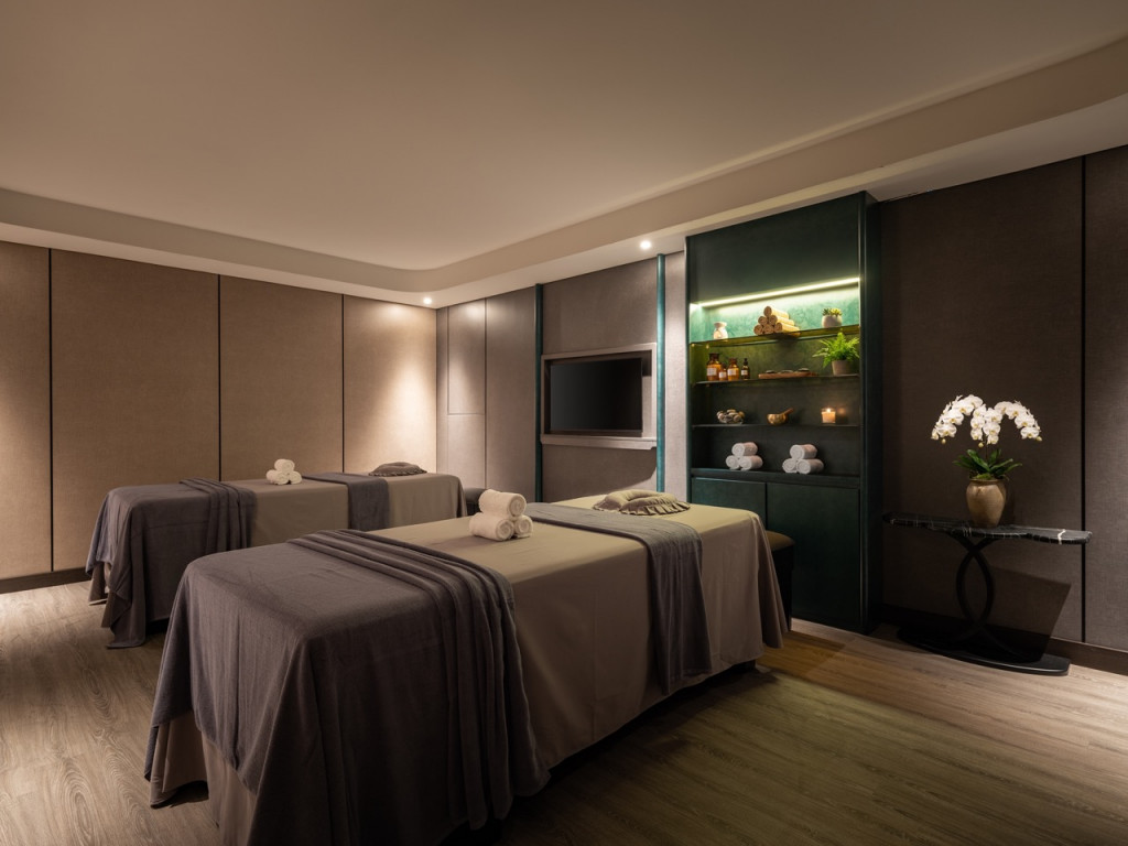 Spa treatment room with three massage tables, neutral linens, rolled towels, and a teal display cabinet with spa products and plants in soft lighting