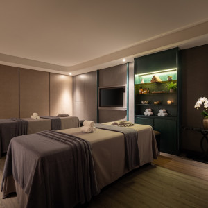 Spa treatment room with three massage tables, neutral linens, rolled towels, and a teal display cabinet with spa products and plants in soft lighting