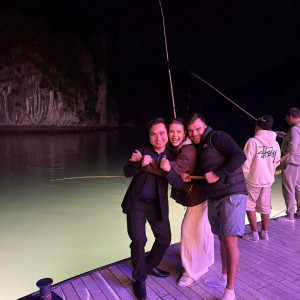 Three friends posing with fists up on a wooden dock at night by a pale-green lake, fishing rods visible in the background.