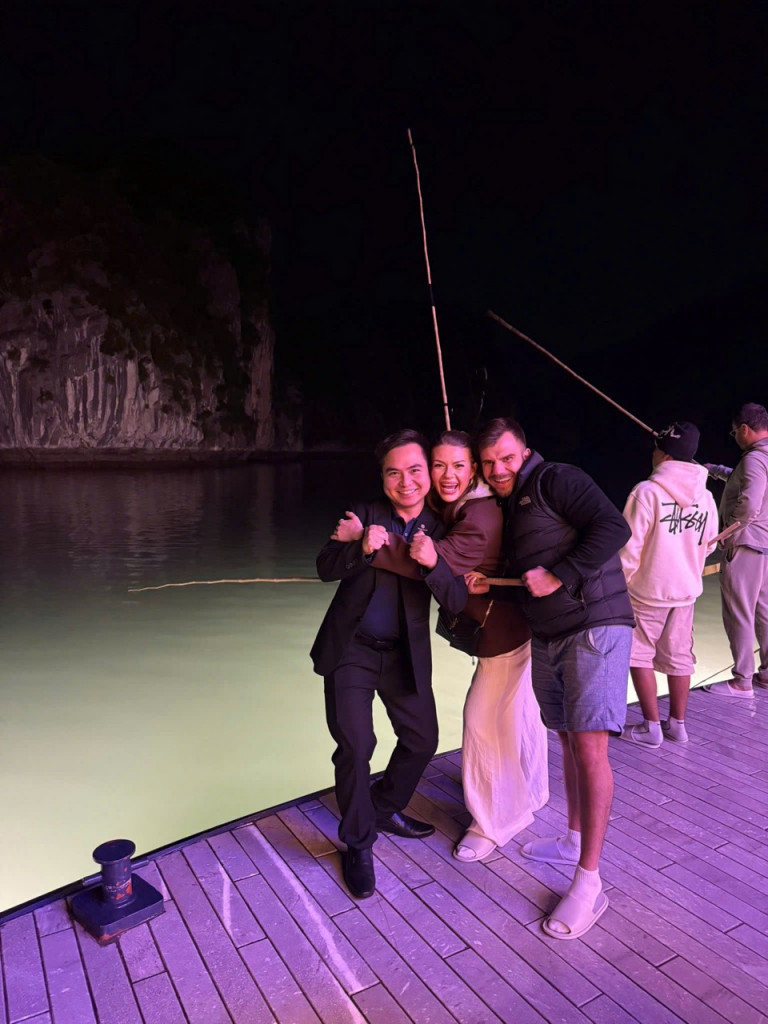 Three friends posing with fists up on a wooden dock at night by a pale-green lake, fishing rods visible in the background.