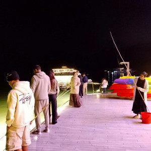Nighttime scene of people lined up along a lit boardwalk with fishing rods, by the water, with boats and colorful kayaks in the background.
