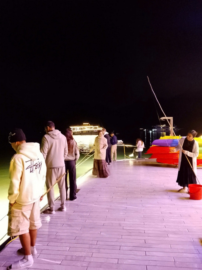 Nighttime scene of people lined up along a lit boardwalk with fishing rods, by the water, with boats and colorful kayaks in the background.