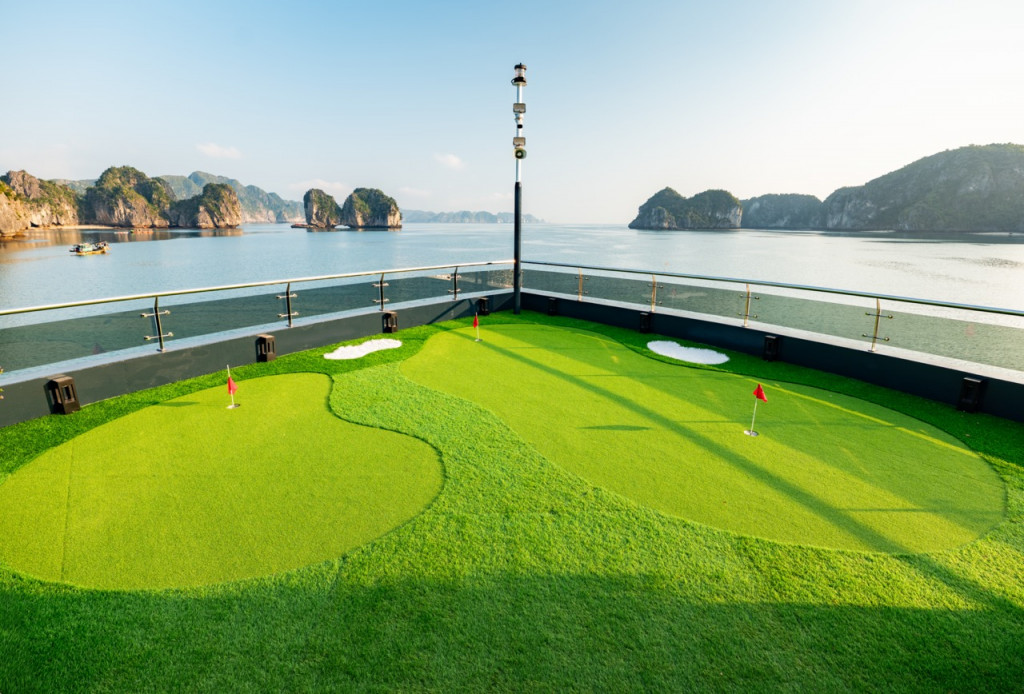Miniature golf putting green on a ship deck with red-hole flags and a calm sea of rocky islets in the background