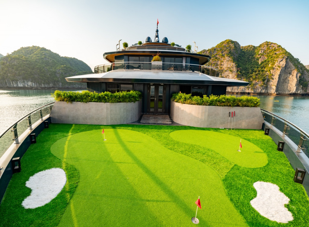 Luxury yacht on calm water with a rooftop deck and a bright artificial turf mini-golf course on the forward deck, surrounded by rocky islands.