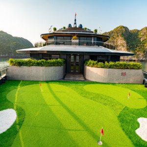 Luxury yacht on calm water with a rooftop deck and a bright artificial turf mini-golf course on the forward deck, surrounded by rocky islands.
