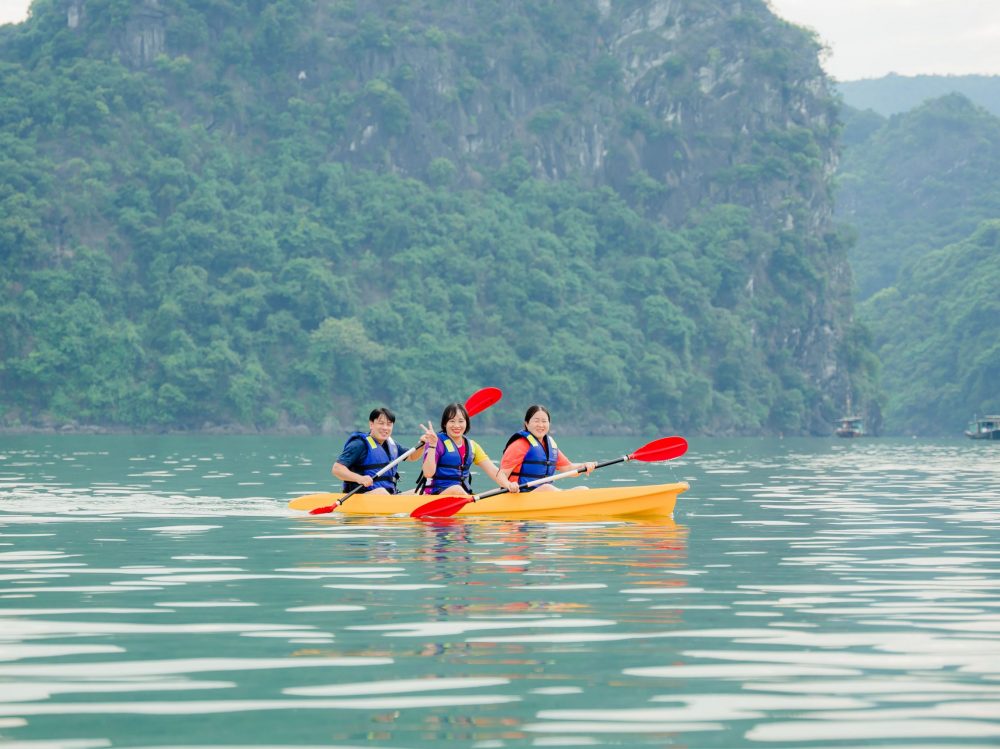 Kayaking scene on Halong Bay featuring three travelers paddling on calm waters with lush green cliff.