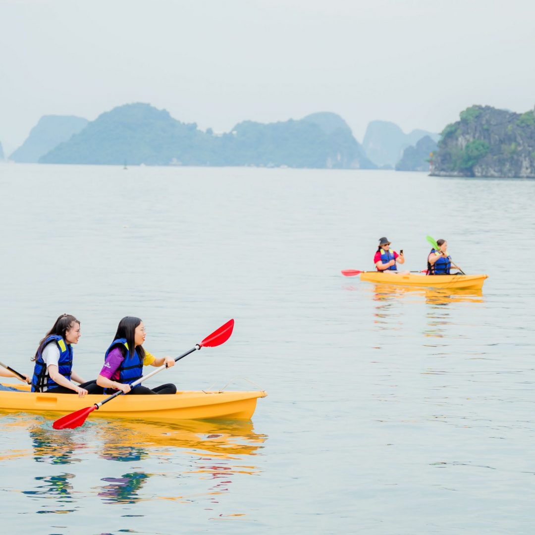 Kayakers paddling on calm Halong Bay waters with limestone islands in the background.